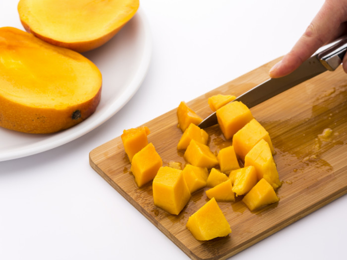 A plate of sliced mango next to a wooden chopping board with small pieces of mango being cut with a knife