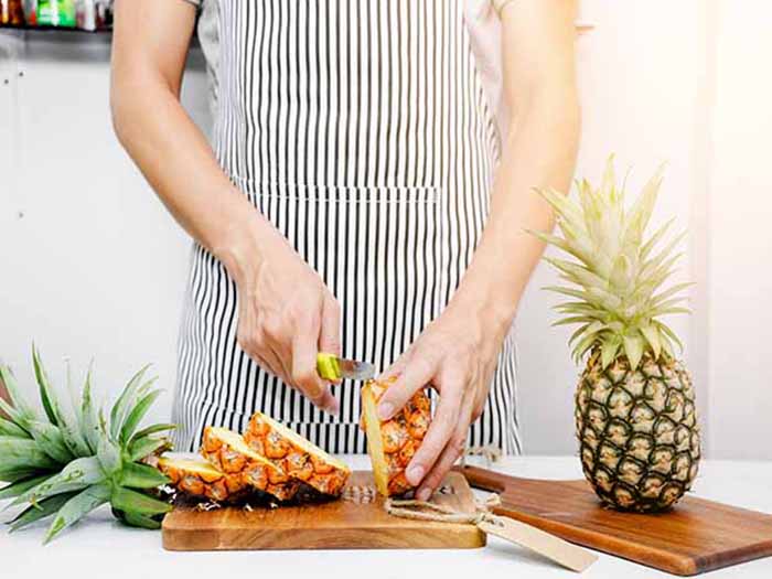 A woman with an apron slicing pineapple on a cutting board with a whole pineapple beside