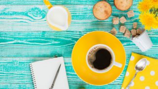 A cup of dandelion coffee next to sugar cubes, a cream jug, muffins, and a journal on a blue counter