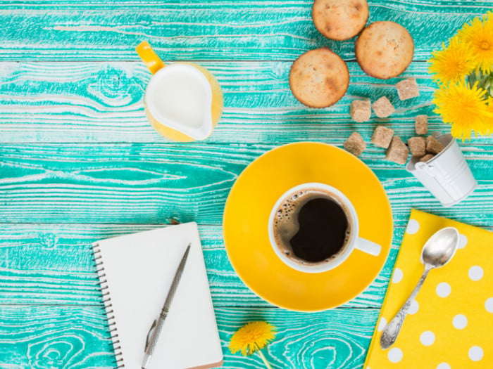 A cup of dandelion coffee next to sugar cubes, a cream jug, muffins, and a journal on a blue counter