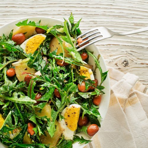 Close up of dandelion leaves, potato, beans and boiled eggs on white plate with a fork