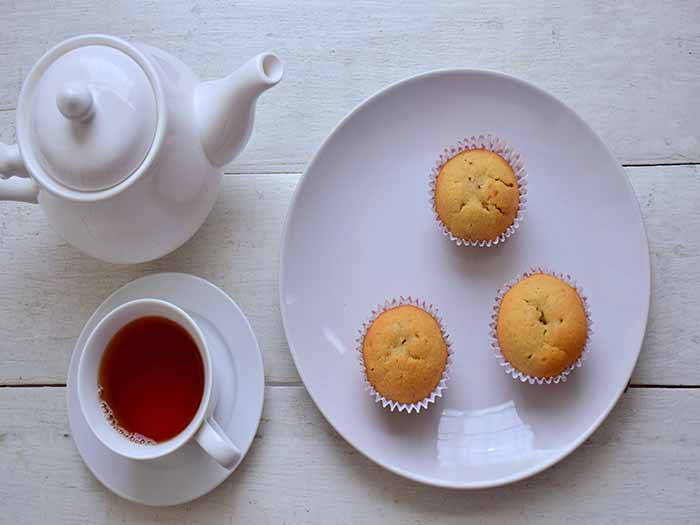 Flatlay picture of a pot of tea, cup and saucer of tea with a plate containing three cupcakes on a white wooden surface