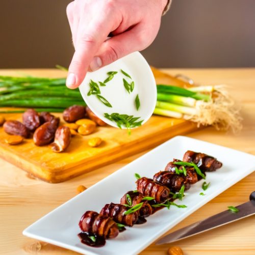 Close up on a hand putting sliced green onions on dates wrapped in bacon