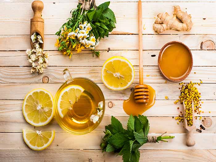 A cup of tea with a lemon slice in it next to dried daisy flowers, honey, and ginger