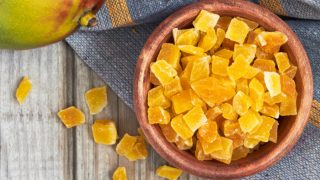 A wooden bowl with sliced dried mango pieces and a whole fresh mango on a wooden table