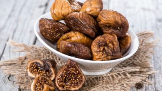 A bowl of dried fig in a white bowl, alongside few more dried figs, placed on a wooden surface.