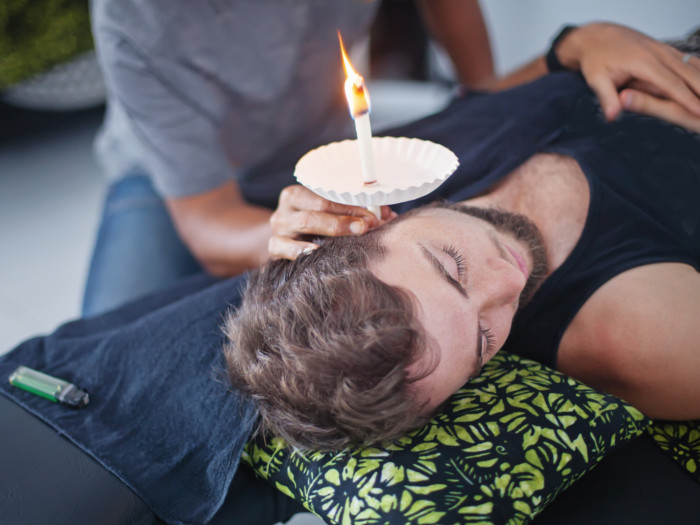 A picture of a man lying down while someone else conducts ear candling on him