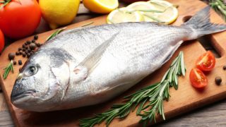 A whole fish on a wooden tray with peppercorns, thyme, half-cut cherry tomato, and other vegetables