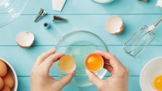 Hand holding eggshells over a bowl, surrounded by a whisk, eggs, piping bag, and top, placed on a light blue wooden surface