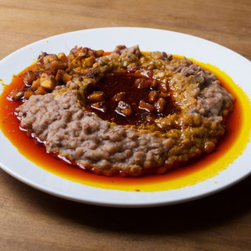 Ewa agoyin plate of mashed beans with diced fried plantain and red palm oil stew on a wooden counter