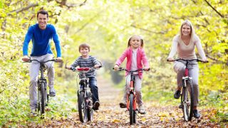 A nuclear family happily cycling in a trail, with autumn leaves in the background