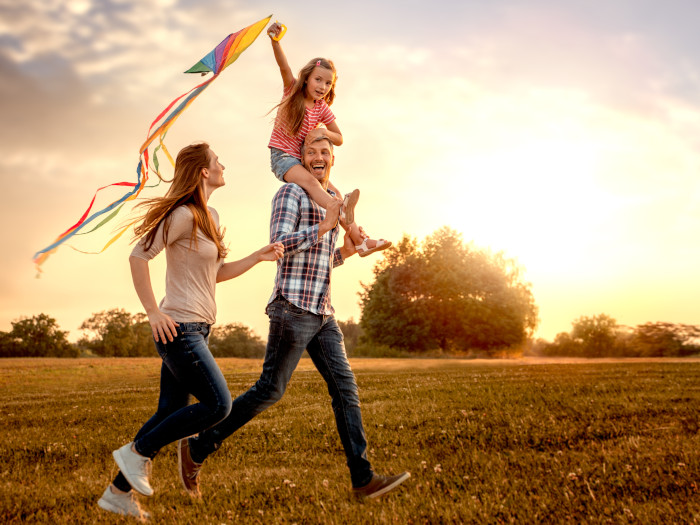 A family of three running on grass with a little girl sitting on a shoulder with a kite in her hand