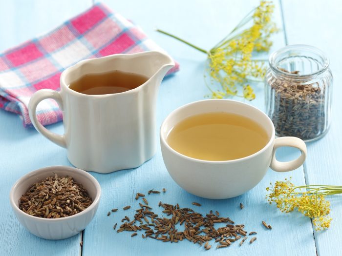 Uma chávena branca e uma jarra cheia de chá de funcho com uma tigela e recipiente de sementes de funcho numa mesa de madeira