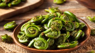 Green fiddlehead ferns in a brown bowl on a jute cloth on wooden table