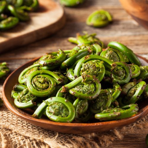 Green fiddlehead ferns in a brown bowl on a jute cloth on wooden table