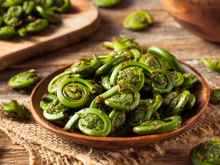 Green fiddlehead ferns in a brown bowl on a jute cloth on wooden table