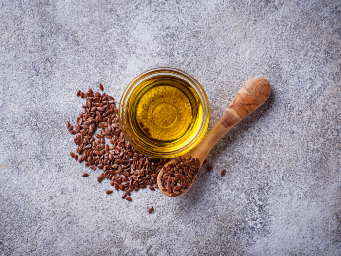 A bowl of flaxseed oil and a wooden spoon filled with flaxseeds on a grey background