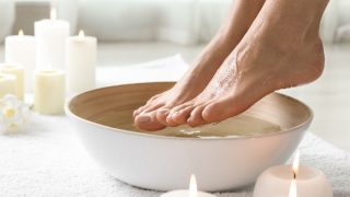 Woman soaking her feet in dish indoors with candles and white towels in the background