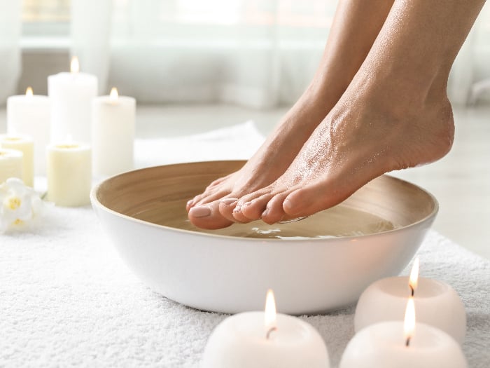 Woman soaking her feet in dish indoors with candles and white towels in the background