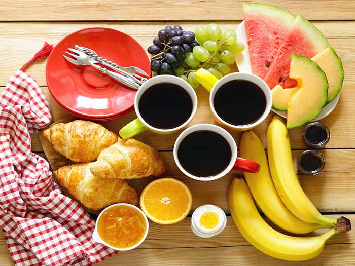 A breakfast platter consisting of bananas, egg, croissants, sliced watermelon, grapes, and black coffee