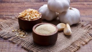 Wooden bowls of garlic powder and sliced garlic with whole and peeled sliced garlic kept on a mat on a wooden table