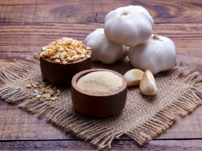 Wooden bowls of garlic powder and sliced garlic with whole and peeled sliced garlic kept on a mat on a wooden table