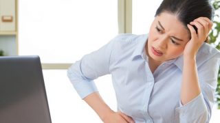 A young woman with stomach pain suffering from gastroenteritis sitting in front of a laptop in an office