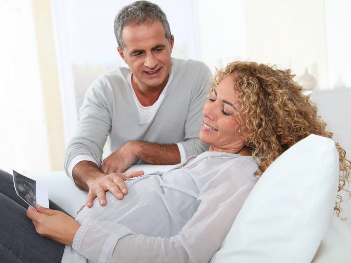 An older pregnant woman smiling while looking at an ultrasound image and an older man with his hands on the woman's stomach.