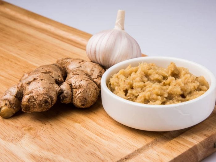 Brown paste in a bowl alongside a ginger knob and a whole garlic bulb on a wooden surface.