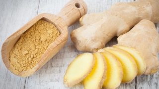 A wooden scoop of ginger powder with whole and sliced gingers on a wooden table
