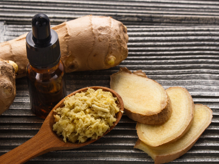A container of ginger root oil, sliced and whole ginger, and a spoon of grated ginger on a table