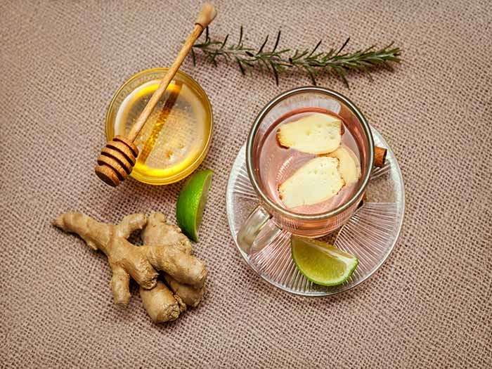 A cup of ginger tea with sliced lemon, a bowl of honey, and ginger on burlap