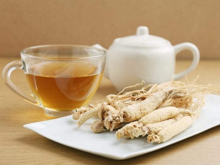 A white cup of ginseng tea, ginseng root, and a kettle on wooden table