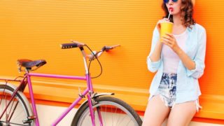 Girl drinking from a yellow paper glass, standing next to a bicycle against a yellow background.
