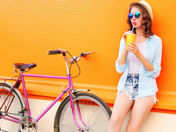 Girl drinking from a yellow paper glass, standing next to a bicycle against a yellow background.