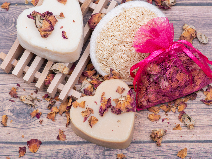 A wooden soap holder with a heart shaped goat milk soap, kept next to a loofah sponge and dried roses on a wooden background.
