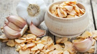A bowl of granulated garlic kept next to garlic, atop a wooden table