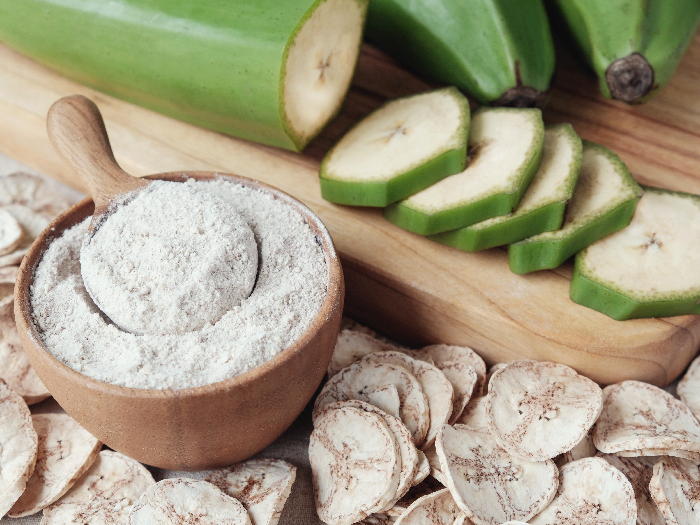 Raw green banana plantain flour in a bowl with sliced plantains and whole plantains in the background