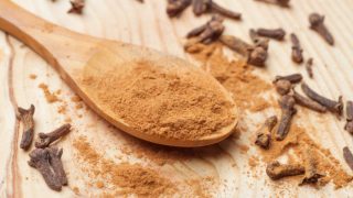 Ground cloves in a wooden spoon next to whole cloves on a wooden surface