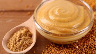 Ground mustard in a spoon next to mustard sauce in a bowl and mustard seeds on the tabletop