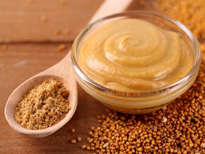 Ground mustard in a spoon next to mustard sauce in a bowl and mustard seeds on the tabletop