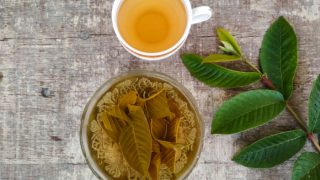 A cup of guava tea, a bowl of containing guava leaves and water, and fresh guava leaves on a wooden table
