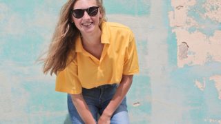 A smiling woman wearing a yellow top, jeans, and black shades, against a blue wall