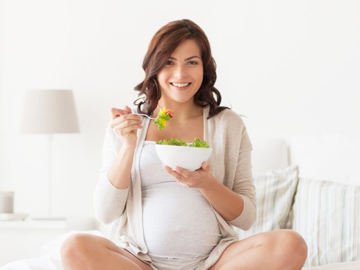 A pregnant woman sitting in a white room with a bowl of salad in her hands, looking into the camera and smiling