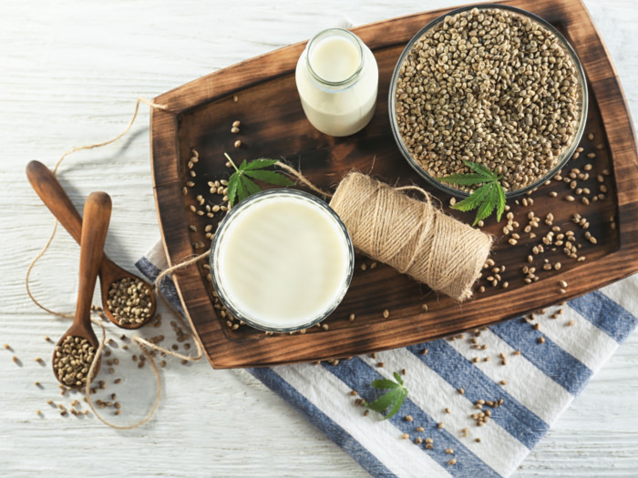 A wooden tray with a bowl of dried hemp seeds, glass and bowl of hemp milk, and a rope and 2 wooden spoons of dried hemp seeds on a wooden table