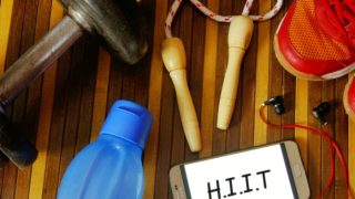 A phone that says HIIT on the screen lying on a table next to earphones, water bottle, dumbbells, shoes, and skipping rope