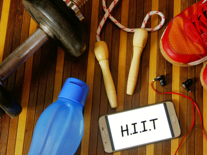 A phone that says HIIT on the screen lying on a table next to earphones, water bottle, dumbbells, shoes, and skipping rope