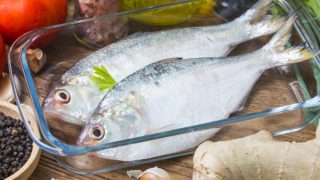 Two fishes in a glass bowl surrounded by ginger, pepper, salt, garlic, oil and tomato on a wooden surface.