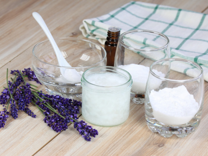 Three bowls containing white powder, essential oil bottle, wax and lavender flowers on a wooden surface.