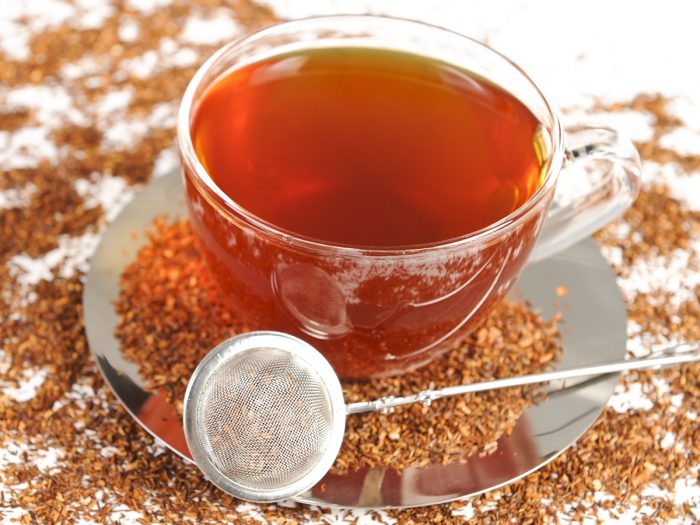 A cup of honeybush tea surrounded by dried honeybush tea leaves and a strainer kept by the side